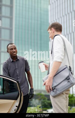 Sourire Noir chauffeur de taxi saluant et ouvrant la porte de la voiture pour le passager caucasien tout en se tenant sur la rue de la ville pendant la journée Banque D'Images
