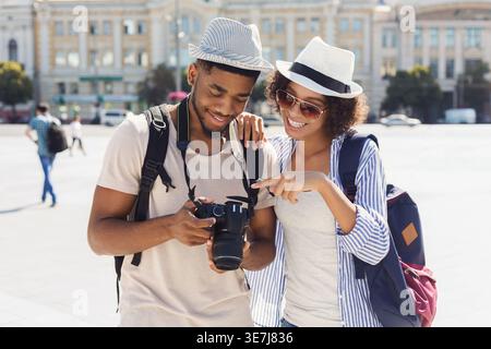 Couple de touristes noirs souriants portant des chapeaux et des lunettes de soleil examinant des photos sur l'appareil photo debout près les uns des autres sur la rue de la ville le jour ensoleillé Banque D'Images