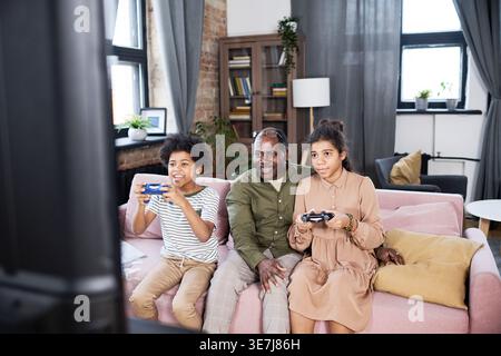 Grand-père noir regardant sur les petits-enfants noirs jouant à des jeux vidéo tout en étant assis sur le canapé ensemble dans le salon à la maison Banque D'Images