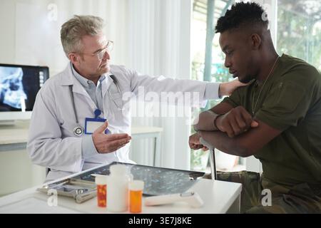 Homme militaire noir sérieux en uniforme militaire avec les bras croisés appuyés sur la canne assise à côté du médecin lui parlant et lui tapant l'épaule dans c... Banque D'Images