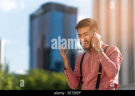 Homme asiatique barbu déçu ayant un appel téléphonique debout dans la rue de la ville pendant la journée Banque D'Images