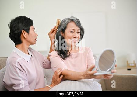 Femme asiatique senior souriante regardant son visage dans le miroir tout en étant assise sur le canapé avec une amie asiatique d'âge moyen peignant ses cheveux avec une brosse... Banque D'Images