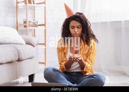 Contrarié femme noire portant chapeau de fête tenant cupcake avec bougie tout en étant assis sur le tapis de sol avec ordinateur portable dans le salon à la maison Banque D'Images
