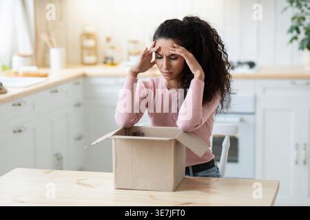Femme frustrée du moyen-Orient touchant la tête regardant la boîte en carton ayant un problème avec son paquet tout en étant assise à table dans la cuisine à la maison Banque D'Images