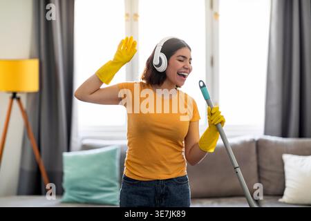 Adolescente hispanique portant des écouteurs et des gants en caoutchouc chantant dans le manche de la serpillière tout en se tenant debout et en nettoyant le salon à la maison Banque D'Images