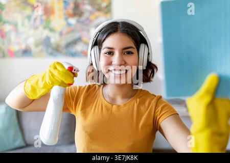 Adolescente hispanique portant des écouteurs et des gants en caoutchouc en utilisant un pulvérisateur et un chiffon dans les fournitures de nettoyage à la maison Banque D'Images
