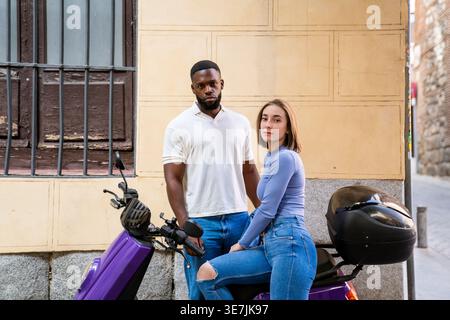 Homme noir sérieux avec la barbe debout à côté de la petite amie caucasienne assis sur le scooter violet sur la rue de la ville pendant la journée Banque D'Images