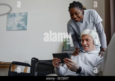 Soignante noire en uniforme tenant les épaules d'un patient d'âge moyen caucasien regardant sur tablette ensemble dans le salon à la maison Banque D'Images