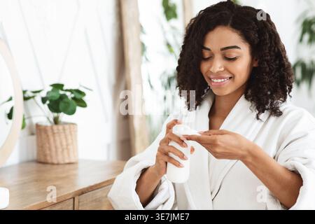 Femme noire en peignoir appliquant le lait nettoyant sur le coton pour enlever le maquillage du visage assis sur la chaise dans la chambre à coucher à la maison Banque D'Images