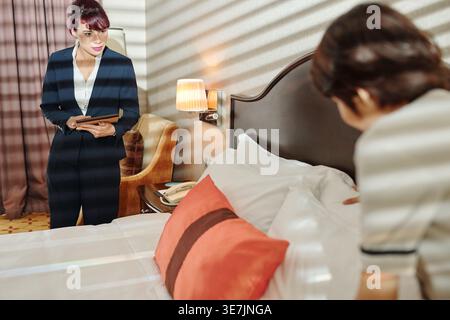 Directrice d'hôtel mixte tenant une tablette regardant une femme de ménage asiatique faire un lit dans la chambre d'hôtel Banque D'Images