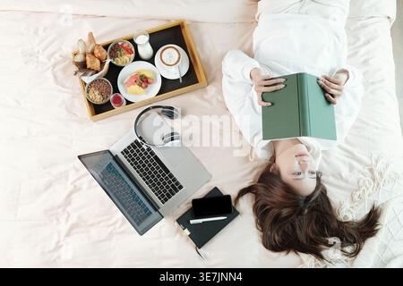 Femme asiatique lisant le livre tout en étant allongée sur le lit près de l'ordinateur portable et plateau de petit déjeuner dans la chambre à la maison Banque D'Images