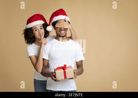 Femme noire portant chapeau de Père Noël faisant silence geste de la main couvrant les yeux de petit ami tenant la boîte-cadeau tout en se tenant debout sur fond marron dans St... Banque D'Images