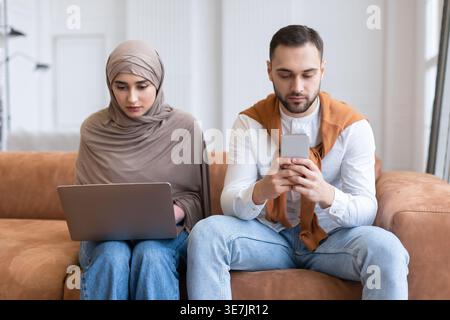 Femme musulmane arabe en hijab et mari caucasien utilisant un ordinateur portable et un smartphone tout en étant assis ensemble sur un canapé dans le salon à la maison Banque D'Images