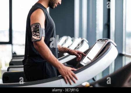 Image recadrée d'un homme noir avec une barbe portant un brassard debout et sélectionnant le mode sur le tapis roulant avant de faire du jogging dans la salle de gym Banque D'Images