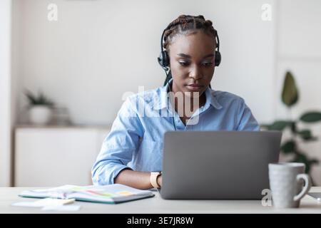Femme d'affaires noire avec des tresses portant des écouteurs travaillant sur ordinateur portable tout en étant assis à table dans le bureau à domicile Banque D'Images