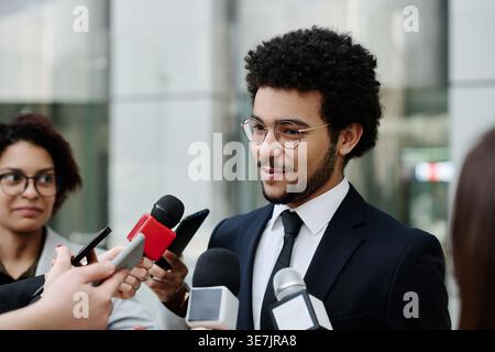 Homme d'affaires de race mixte en costume et lunettes parlant aux journalistes donnant une interview à la conférence d'affaires tout en se tenant debout à l'extérieur du bureau bui... Banque D'Images