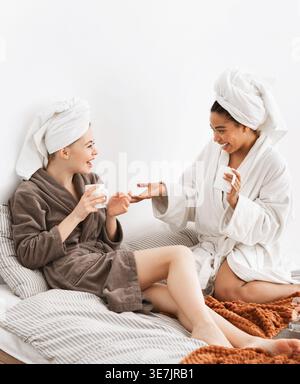 Joyeuses amies noires et caucasiennes portant des peignoirs avec des têtes enveloppées tenant des tasses à café et parlant les unes avec les autres tout en restant assis toge... Banque D'Images