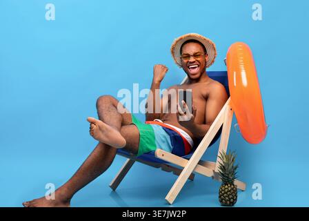 Homme noir portant des lunettes et chapeau de paille avec poing serré tenant smartphone assis avec les jambes croisées sur chaise longue avec anneau gonflable aga... Banque D'Images