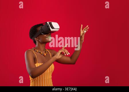 Femme noire souriante avec les cheveux courts portant un casque de réalité virtuelle tout en se tenant devant un fond rouge en studio Banque D'Images