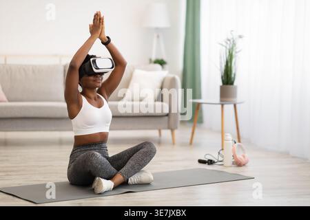 Femme noire avec des vêtements de sport bouclés et casque VR méditant sur tapis dans le salon à la maison Banque D'Images