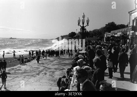 Crimée, Gurzuf ou Yalta, 1968. Une vague de mer massive s'écrase dramatiquement contre le remblai de béton pavé, pulvérisant de l'eau de mer dans les airs. Une grande foule de curieux vacanciers d'été se réunit le long de la magnifique promenade du bord de mer pour regarder le puissant surf orageux sous le soleil chaud et lumineux. Cette photographie de rue d'archives très dynamique capture magnifiquement l'énergie brute de la nature côtière et les loisirs de plein air populaires dans une station thermale soviétique classique, reflétant la vie quotidienne heureuse et paisible avant la guerre. Banque D'Images