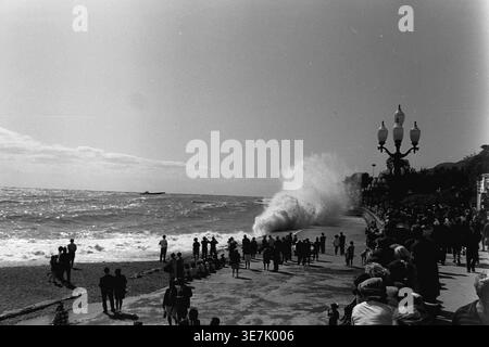 Crimée, Gurzuf ou Yalta, 1968. Une vague de mer massive s'écrase dramatiquement contre le remblai de béton pavé, pulvérisant de l'eau de mer dans les airs. Une grande foule de curieux vacanciers d'été se réunit le long de la magnifique promenade du bord de mer pour regarder le puissant surf orageux sous le soleil chaud et lumineux. Cette photographie de rue d'archives très dynamique capture magnifiquement l'énergie brute de la nature côtière et les loisirs de plein air populaires dans une station thermale soviétique classique, reflétant la vie quotidienne heureuse et paisible avant la guerre. Banque D'Images