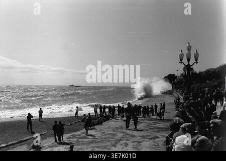Crimée, Gurzuf ou Yalta, 1968. Une vague de mer massive s'écrase dramatiquement contre le remblai de béton pavé, pulvérisant de l'eau de mer dans les airs. Une grande foule de curieux vacanciers d'été se réunit le long de la magnifique promenade du bord de mer pour regarder le puissant surf orageux sous le soleil chaud et lumineux. Cette photographie de rue d'archives très dynamique capture magnifiquement l'énergie brute de la nature côtière et les loisirs de plein air populaires dans une station thermale soviétique classique, reflétant la vie quotidienne heureuse et paisible avant la guerre. Banque D'Images