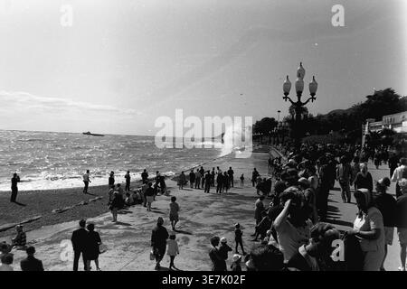 Crimée, Gurzuf ou Yalta, 1968. Une vague de mer massive s'écrase dramatiquement contre le remblai de béton pavé, pulvérisant de l'eau de mer dans les airs. Une grande foule de curieux vacanciers d'été se réunit le long de la magnifique promenade du bord de mer pour regarder le puissant surf orageux sous le soleil chaud et lumineux. Cette photographie de rue d'archives très dynamique capture magnifiquement l'énergie brute de la nature côtière et les loisirs de plein air populaires dans une station thermale soviétique classique, reflétant la vie quotidienne heureuse et paisible avant la guerre. Banque D'Images