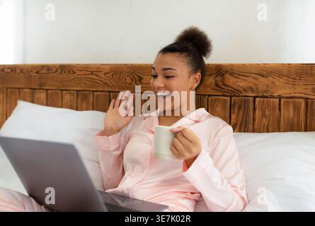 Femelle noire en pyjama tenant une tasse à café agitant la main ayant un appel vidéo à l'aide d'un ordinateur portable assis sur le lit à la maison Banque D'Images