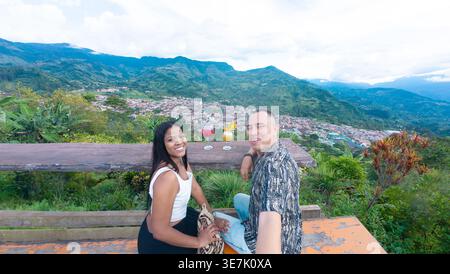 Couple noir et hispanique prenant selfie tout en étant assis à une table en bois avec des cocktails en face du paysage de montagne Banque D'Images