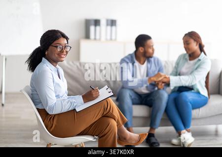 Psychothérapeute femme noire avec des lunettes tenant le presse-papiers tout en étant assis devant le patient de couple tenant les mains de l'autre sur le canapé pendant t... Banque D'Images