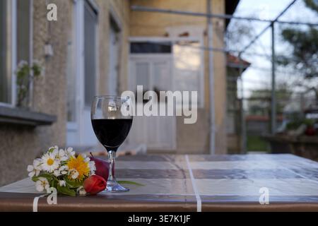 Nature morte en plein air avec un verre de vin rouge, une pomme mûre et des fleurs printanières sur une table réfléchissante, avec une maison et des arbres légèrement flous. 2026 Banque D'Images