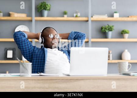 Homme d'affaires noir portant des lunettes et des écouteurs regardant loin assis à la table avec les mains derrière la tête dans le bureau Banque D'Images