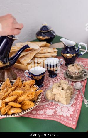 La main verse le café d'une théière vintage bleu cobalt dans une tasse. Pâtisseries orientales traditionnelles baklava et halva. Nourriture sucrée. Banque D'Images