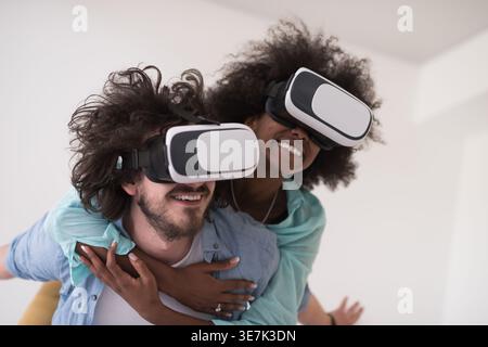 Couple noir et caucasien portant des casques VR ayant un plaisir piggyback équitation et expérimentant la réalité virtuelle sur fond blanc en studio Banque D'Images