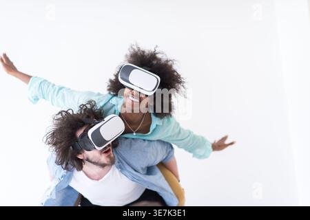 Couple noir et caucasien portant des casques VR ayant un plaisir piggyback équitation et expérimentant la réalité virtuelle sur fond blanc en studio Banque D'Images