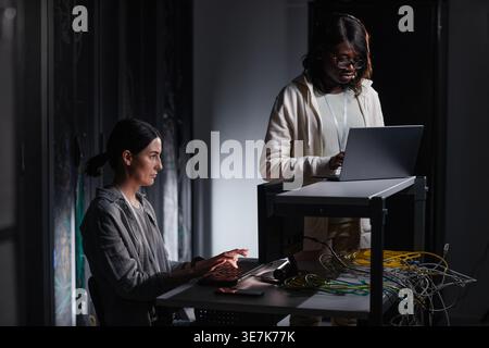 Ingénieur INFORMATIQUE féminin noir portant des lunettes debout et à l'aide d'un ordinateur portable près d'une collègue féminine caucasienne travaillant sur l'ordinateur assis à table dans servir... Banque D'Images