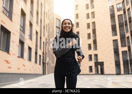 Femme musulmane du moyen-Orient souriante avec tracker de fitness portant hijab et vêtements de sport courant dans la rue pendant la journée Banque D'Images