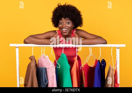 Femme noire avec afro montrant les pouces vers le haut s'appuyant sur le porte-vêtements tout en se tenant debout sur fond jaune dans le studio Banque D'Images