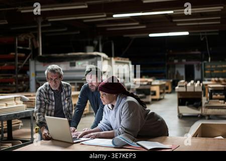 Femme noire debout avec des charpentiers caucasiens utilisant un ordinateur portable dans un magasin de menuiserie Banque D'Images
