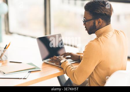 Homme noir portant des lunettes en utilisant un ordinateur portable assis à table avec des cahiers dans le bureau Banque D'Images
