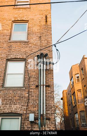 En milieu urbain, des fils électriques longent le côté d’un bâtiment en briques avec plusieurs fenêtres. La scène montre un ciel clair et des briques bien usées un Banque D'Images