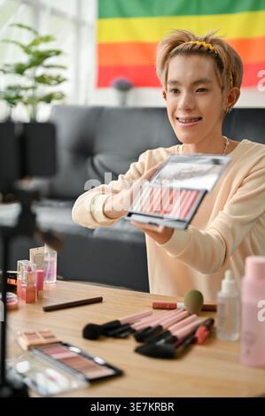 Homme gay asiatique souriant montrant sa palette de fards à paupières sur la caméra tout en enregistrant le tutoriel de maquillage assis sur le sol près du canapé à la maison Banque D'Images
