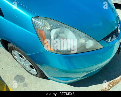 Une voiture bleue est garée dans un lot sous le soleil. Le phare avant présente de la saleté et de la poussière. Le cadre est dans une zone dégagée avec d'autres voitures à proximité. Banque D'Images