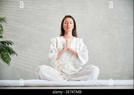 Belle femme en robe blanche assise sur tapis en position lotus avant massage thaïlandais, pratiquant le yoga, montrant signe namaste. Banque D'Images