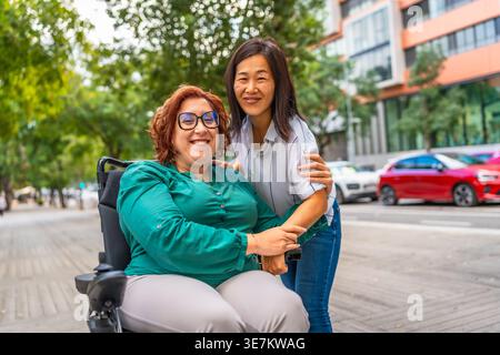 Femme chinoise d'âge moyen debout à côté d'une femme caucasienne avec un handicap dans tout en chaise tout en ville pendant la journée Banque D'Images