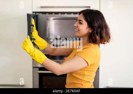 Adolescente hispanique portant des gants en caoutchouc en utilisant un chiffon dans le micro-ondes de nettoyage tout en se tenant debout dans la cuisine à la maison Banque D'Images