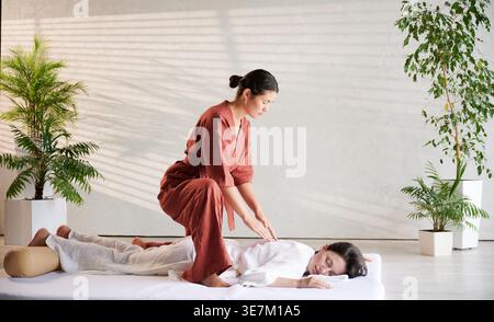 Masseuse faisant massage yoga thaïlandais. Thérapeute de femme faisant le traitement de massage traditionnel pour la patiente féminine à l'intérieur. Client allongé sur tapis, méditant, profitant d'un massage des mains dans une atmosphère calme. Banque D'Images