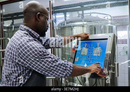 Homme chauve noir dans le tablier de la machine utilisant le moniteur d'ordinateur à écran tactile debout en usine Banque D'Images