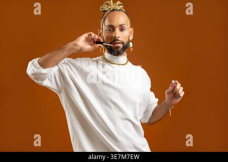 Homme gay brésilien sérieux avec les cheveux tressés portant un pull blanc col roulé avec maquillage coloré des yeux appliquant le rouge à lèvres debout contre l'orange BA... Banque D'Images